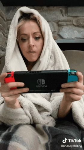 A Woman Wrapped In A Blanket Is Playing A Game On A Nintendo Switch GIF