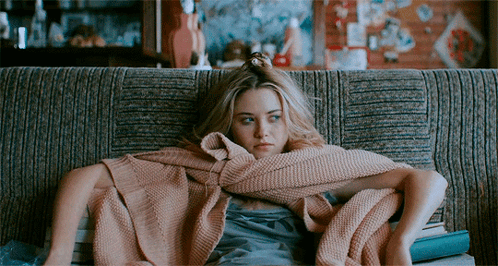 A Woman Wrapped In A Pink Blanket Sits On A Couch With Books GIF