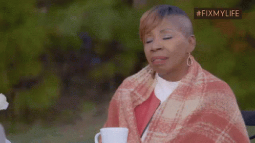 A Woman Wrapped In A Plaid Blanket Is Sitting At A Table With A Cup Of Coffee . GIF