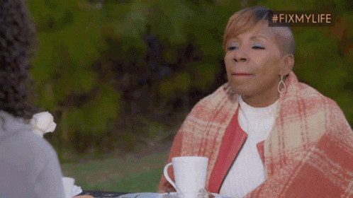A Woman Wrapped In A Plaid Blanket Sits At A Table With A Cup Of Coffee In Front Of Her GIF