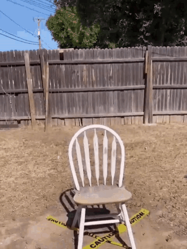 A Wooden Chair Sits In The Dirt In Front Of A Fence With A Yellow Caution Tape Around It GIF