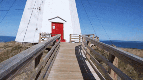 A Wooden Walkway Leads To A White Building With A Red Door And A Sign That Says No Dogs GIF