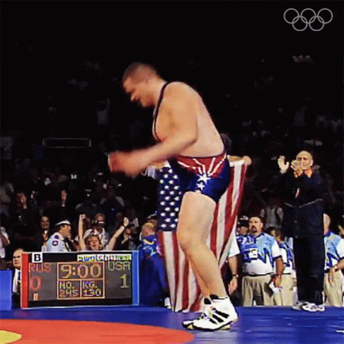 A Wrestler Holds An American Flag In Front Of A Scoreboard That Says " Bud Light " On It GIF