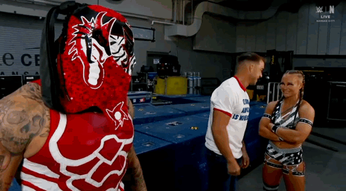 A Wrestler In A Red And White Outfit Is Talking To A Man And A Woman In A Room . GIF