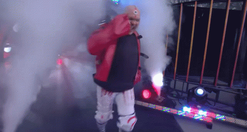a wrestler in a red jacket and white pants is walking through smoke