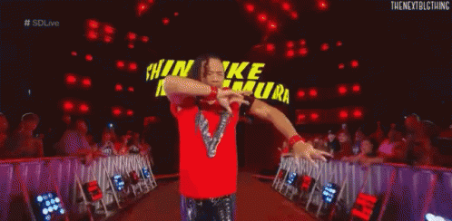 a wrestler in a red shirt is kneeling down in front of a large sign that says shinsuke nakamura .