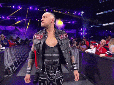 A Wrestler In A Studded Vest Stands In Front Of A Crowd At A Wrestling Match GIF