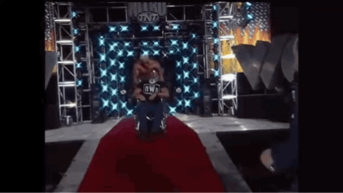 A Wrestler In A Wheelchair Is Sitting On A Red Carpet On A Stage . GIF