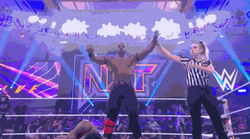 A Wrestler Is Being Congratulated By A Referee In Front Of A Sign That Says Nxt GIF