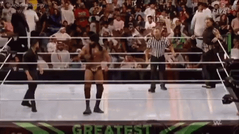 A Wrestler Is Dancing In A Wrestling Ring With A Referee In Front Of A Crowd . GIF