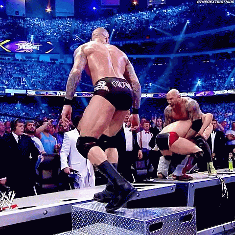 A Wrestler Is Jumping Over A Metal Box While Another Wrestler Is Kneeling Down . GIF