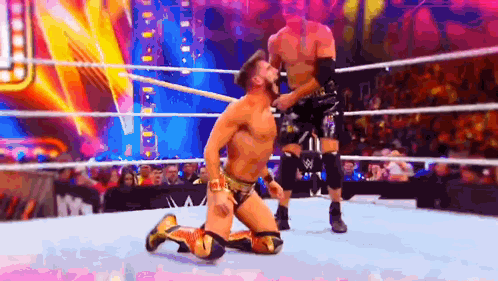a wrestler is kneeling down in a wrestling ring while another wrestler is standing behind him .