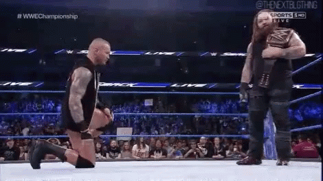 A Wrestler Is Kneeling Down In A Wrestling Ring While Another Wrestler Stands Behind Him . GIF