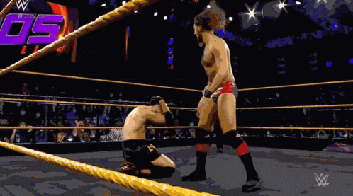 A Wrestler Is Kneeling Down In A Wrestling Ring While Another Wrestler Stands In The Corner GIF