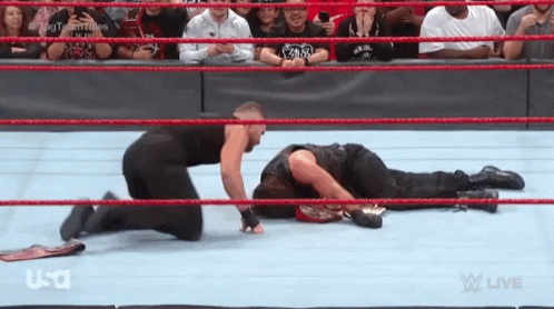 A Wrestler Is Kneeling Down Next To Another Wrestler Who Is Laying On The Floor . GIF