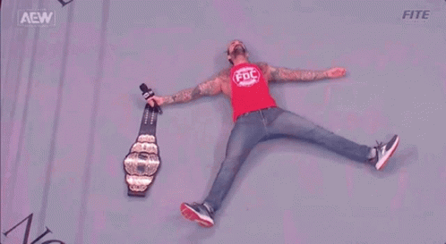 A Wrestler Is Laying On The Floor With A Microphone And A Championship Belt . GIF