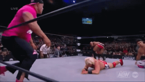 A Wrestler Is Laying On The Ground In A Wrestling Ring While A Man In A Pink Shirt Looks On . GIF