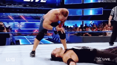 A Wrestler Is Laying On The Ground While Another Wrestler Is Standing On Top Of Him . GIF