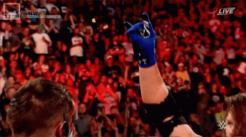 A Wrestler Is Raising His Hand In The Air In Front Of A Crowd While Wearing A Blue Glove . GIF