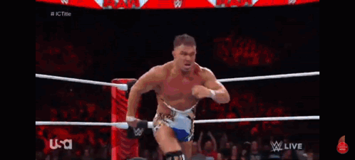 A Wrestler Is Standing In The Middle Of A Wrestling Ring While Another Wrestler Is Kneeling Down . GIF