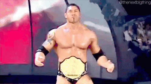 A Wrestler Is Wearing A Championship Belt And Standing On A Stage With His Fist In The Air . GIF