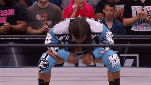 A Wrestler Wearing Knee Pads With Ac On Them Is Kneeling Down In Front Of A Crowd GIF