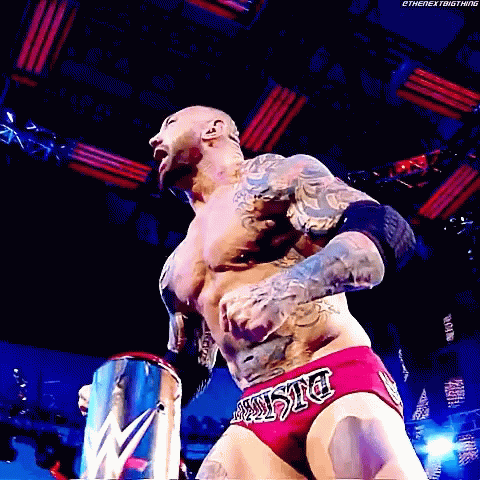 A Wrestler With A Tattoo On His Chest Is Wearing A Pair Of Red Trunks With The Word Justice On The Side GIF