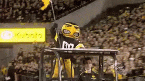 A Yellow And Black Mascot Is Sitting On Top Of A Tractor Holding A Flag . GIF