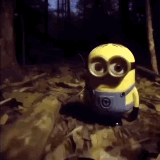 a yellow minion with big eyes is standing in a dark forest .