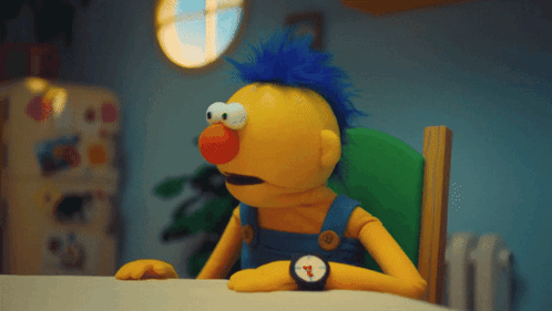 A Yellow Puppet With A Blue Mohawk Sits At A Table GIF