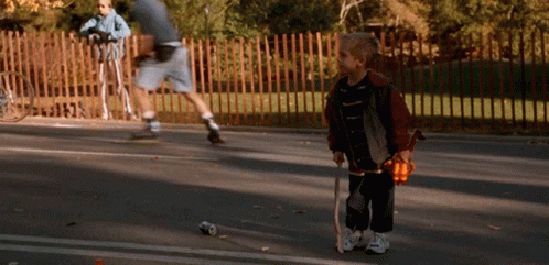 A Young Boy Holding A Cane Stands On The Side Of A Road GIF