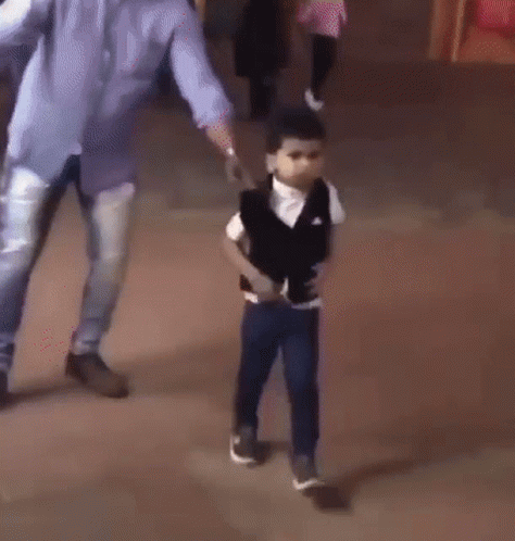 A Young Boy In A Black Vest And White Shirt Is Standing On A Sidewalk . GIF