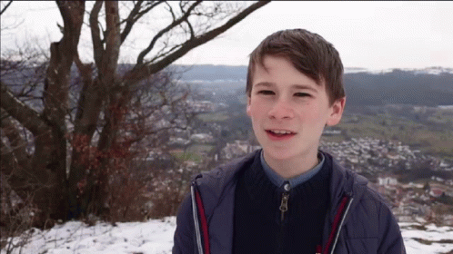 A Young Boy In A Blue Jacket Is Smiling In Front Of A Snowy Landscape GIF
