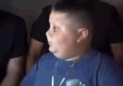 A Young Boy In A Blue Shirt Is Making A Funny Face While Sitting In A Dark Room . GIF
