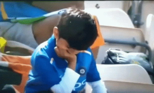 A Young Boy In A Blue Shirt Is Sitting In A Stadium With His Hand On His Face . GIF