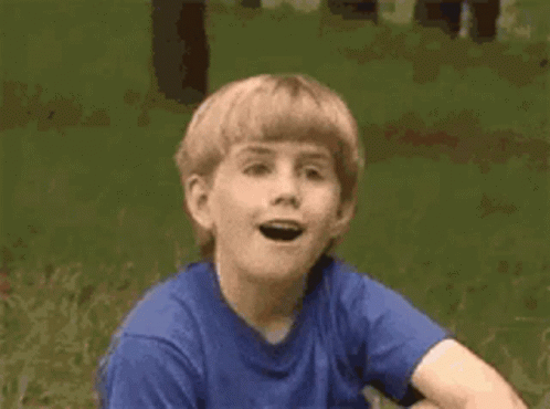 A Young Boy In A Blue Shirt Is Sitting In The Grass . GIF