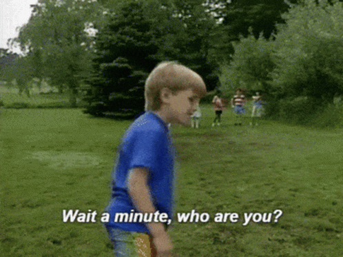 A Young Boy In A Blue Shirt Is Standing In A Grassy Field And Says " Wait A Minute Who Are You " GIF