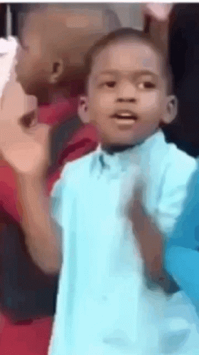 A Young Boy In A Blue Shirt Is Standing In Front Of A Group Of People . GIF
