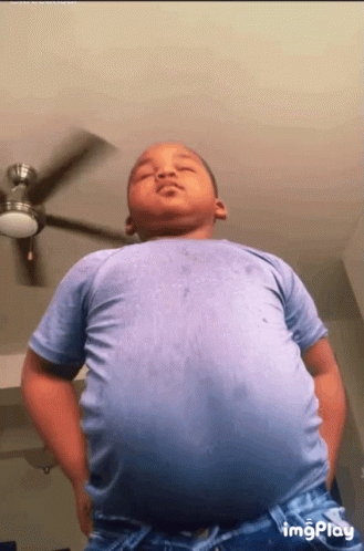 A Young Boy In A Blue Shirt Is Standing Under A Ceiling Fan GIF