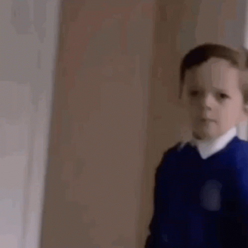 A Young Boy In A Blue Sweater And White Shirt Is Standing In A Room . GIF