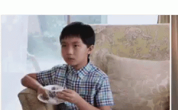 A Young Boy In A Plaid Shirt Is Sitting On A Couch Holding A Cup And Saucer . GIF