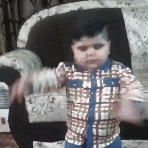 A Young Boy In A Plaid Shirt Is Standing In Front Of A Couch . GIF