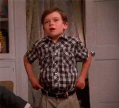 A Young Boy In A Plaid Shirt Is Standing In Front Of A Door Holding A Spoon And Saying `` Fashion '' . GIF