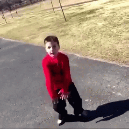 A Young Boy In A Red Shirt And Black Pants Is Standing On A Sidewalk . GIF