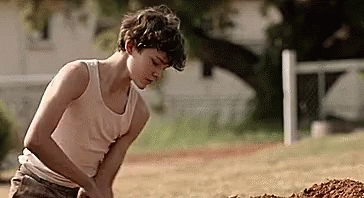 A Young Boy In A Tank Top Is Digging In The Dirt In A Field . GIF