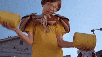 A Young Boy In A Yellow Shirt Is Eating Noodles From A Bowl While Holding Two Yellow Containers . GIF