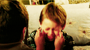 A Young Boy Is Covering His Face With His Hands While Sitting On A Bed . GIF