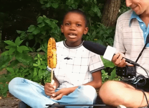 A Young Boy Is Holding A Corn On The Cob And Talking Into A Microphone GIF