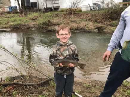 A Young Boy Is Holding A Fish In His Hands In Front Of A Stream . GIF