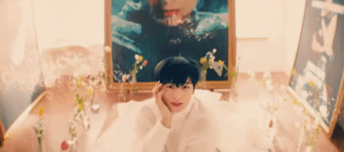 A Young Boy Is Laying In A Bathtub With Flowers And A Picture Of A Woman In The Background . GIF